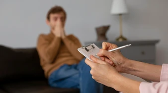 Mãos de uma psicóloga em primeiro plano fazendo anotações em uma prancheta durante sessão de avaliação psicológica em Brasília. Ao fundo, desfocado, um paciente homem está sentado no sofá do consultório.