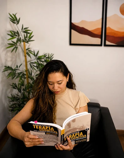 Neuropsicóloga Sara Alves sentada em uma poltrona preta lendo o livro A Prática Clínica de Terapia Cognitiva com Crianças e Adolescentes em seu consultório em Brasília.